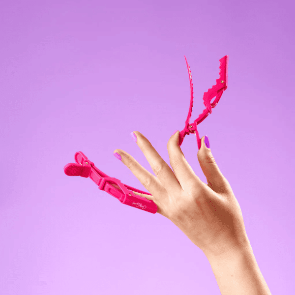 Hand met roze nagellak die een roze Curly Secret alligator hair clip vasthoudt tegen een paarse achtergrond.