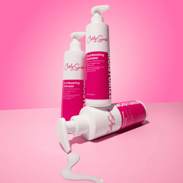 Drie flessen Curly Secret Curl Boosting Activator speels opgesteld, waarvan één fles product uit de pomp op een roze oppervlak laat lopen.