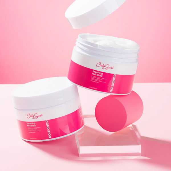 Curly Secret Repairing Hair Mask – open pot haarmasker met romige textuur, roze verpakking, gefotografeerd tegen een roze achtergrond.