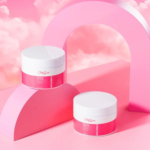 Twee potten Curly Secret Repairing Hair Mask – roze en wit verpakkingsdesign, gepresenteerd in een speelse roze setting met boogvormen.