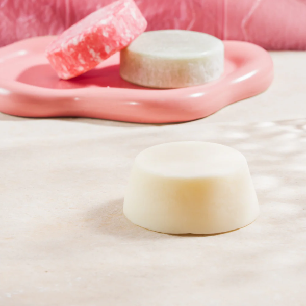 Crème-witte conditioner bar van Curly Secret in een roze schelp, met roze shampoo bars op de achtergrond.