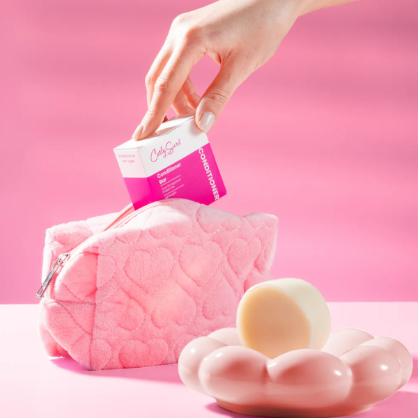 Hand plaatst een Curly Secret Conditioner Bar verpakking in een roze toilettas, met de bar zelf op een roze schaaltje ervoor.