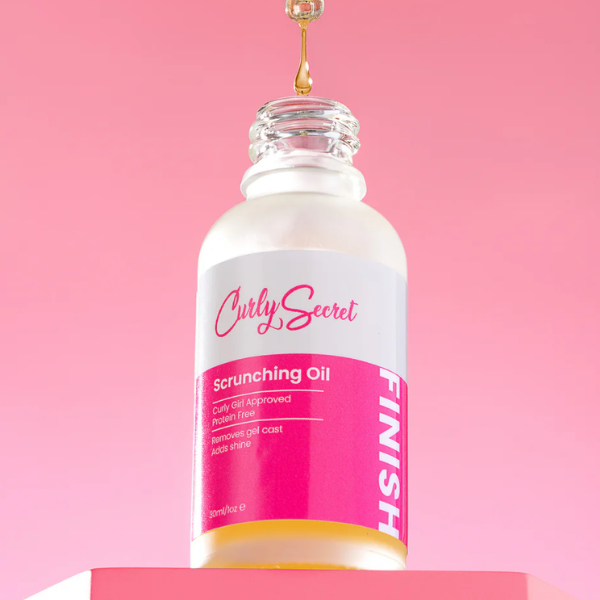 Flesje Curly Secret Scrunching Oil met een pipet waar olie uit druppelt, tegen een roze achtergrond.