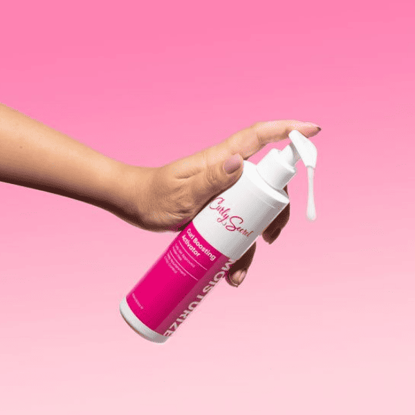 Hand die een fles Curly Secret Curl Boosting Activator vasthoudt tegen een roze achtergrond, waarbij product uit de pomp wordt gedoseerd.