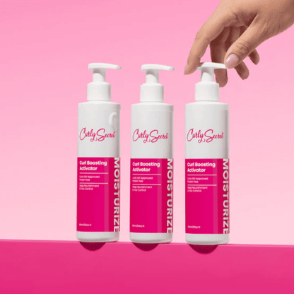 Drie flessen Curly Secret Curl Boosting Activator naast elkaar, met een hand die de pomp van een fles indrukt, op een roze achtergrond.
