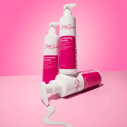 Drie flessen Curly Secret Curl Boosting Activator speels opgesteld, waarvan één fles product uit de pomp op een roze oppervlak laat lopen.