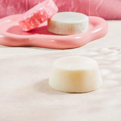 Crème-witte conditioner bar van Curly Secret in een roze schelp, met roze shampoo bars op de achtergrond.