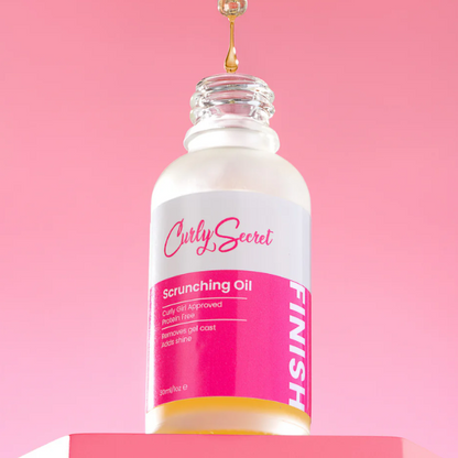 Flesje Curly Secret Scrunching Oil met een pipet waar olie uit druppelt, tegen een roze achtergrond.